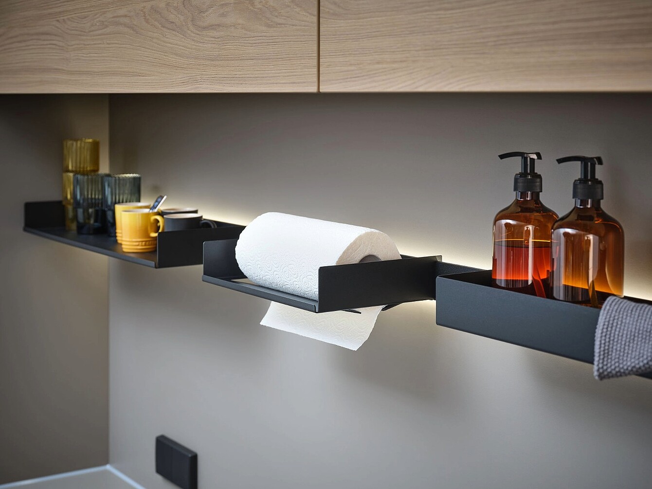 Wall-mounted black metal shelves holding glassware, a yellow cup, a paper towel roll, two amber pump bottles, and a gray cloth under wooden cabinets
