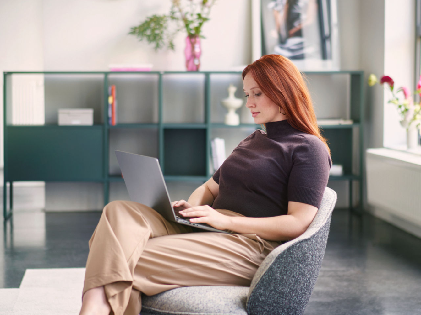 Femme aux cheveux roux, assise détendue dans un fauteuil gris, travaillant sur un ordinateur portable dans une pièce moderne avec des étagères et des plantes en arrière-plan