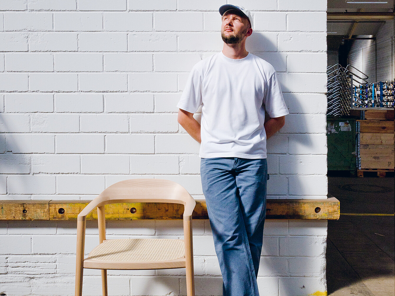 Hombre con camisa blanca informal y pantalones azules de pie contra una pared de ladrillo blanco junto a una silla de madera light se:café diseñada por Konstantin Thomas, destacando su artesanía duradera y cómoda. 