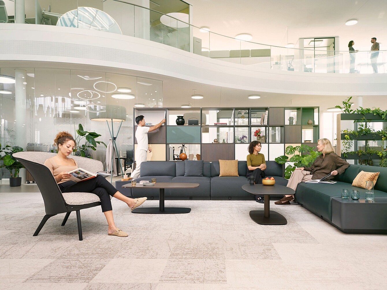 Moderna zona de biblioteca abierta con grupos de asientos, estanterías y personas leyendo, conversando y utilizando una pizarra blanca.