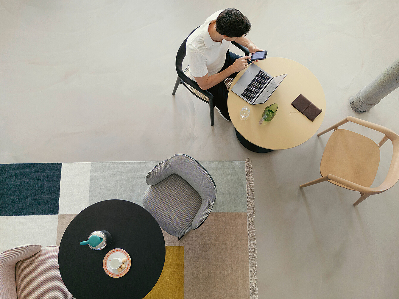Persona seduta a un tavolo rotondo in una moderna biblioteca d’ufficio, che utilizza uno smartphone e un laptop, circondata da sedie e un tappeto, che illustra un rifugio tranquillo per lavorare con concentrazione.