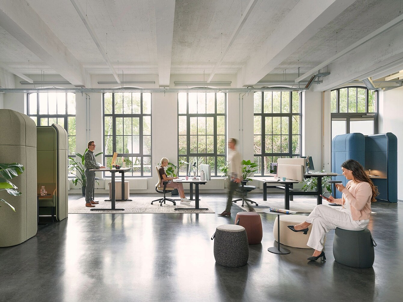 Espace de bureau moderne avec de grandes fenêtres, des bureaux réglables, des modules d’assise et des personnes travaillant ou assises dans un environnement lumineux et ouvert