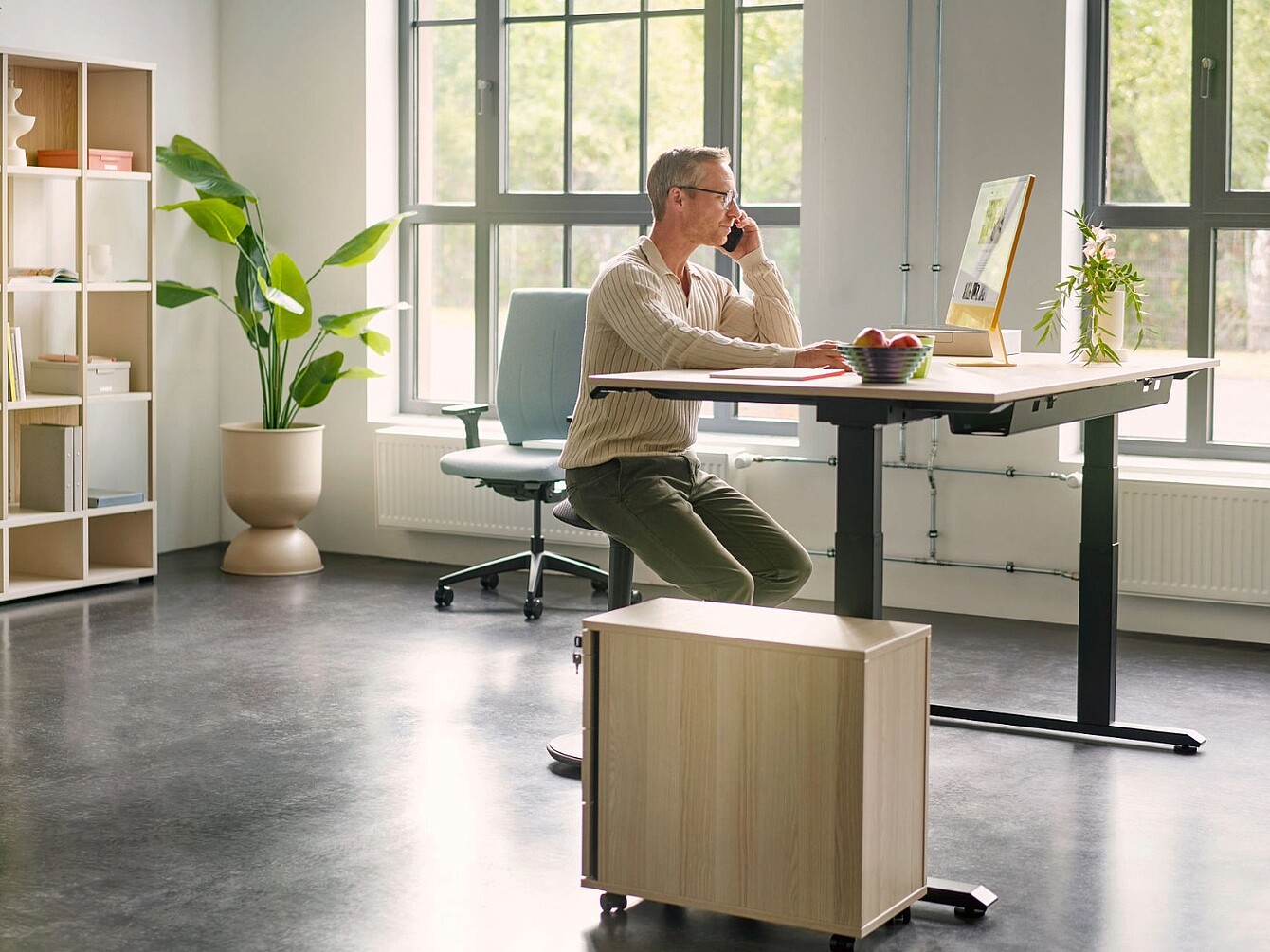 Personne assise sur un tabouret se:fit devant un bureau réglable en hauteur, dans un bureau lumineux doté de grandes fenêtres et de plantes