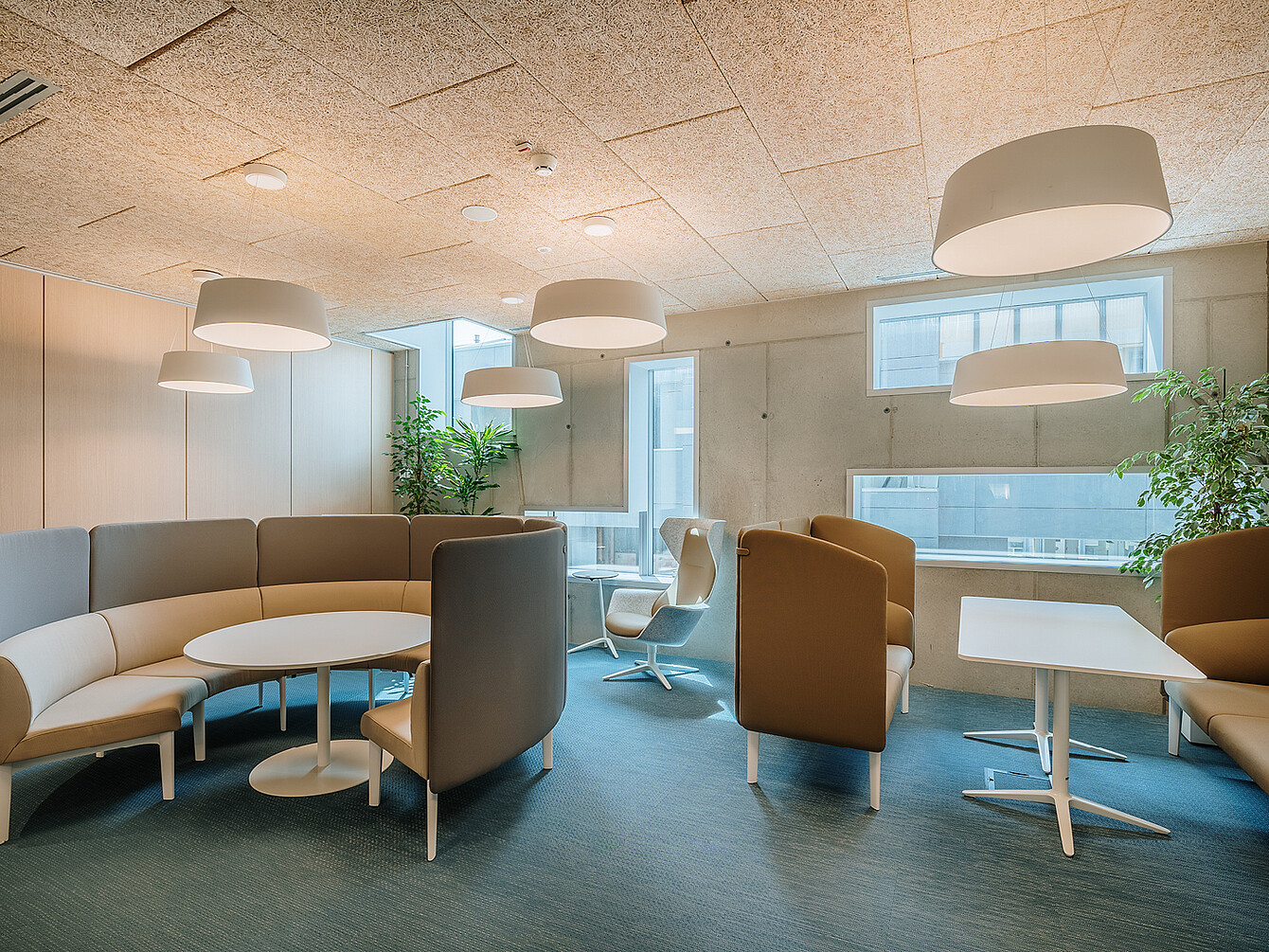 Area lounge dell’ufficio con sedute curve e dallo schienale alto attorno a tavoli rotondi e rettangolari sotto grandi lampade a soffitto e vicino a finestre con piante