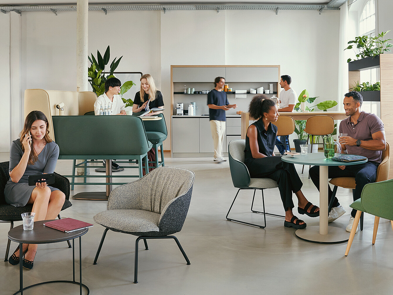 Un Work Café moderno con diferentes disposiciones de asientos. Las personas conversan, algunas utilizan el teléfono. Se ven plantas verdes y, al fondo, hay una pequeña cocina.