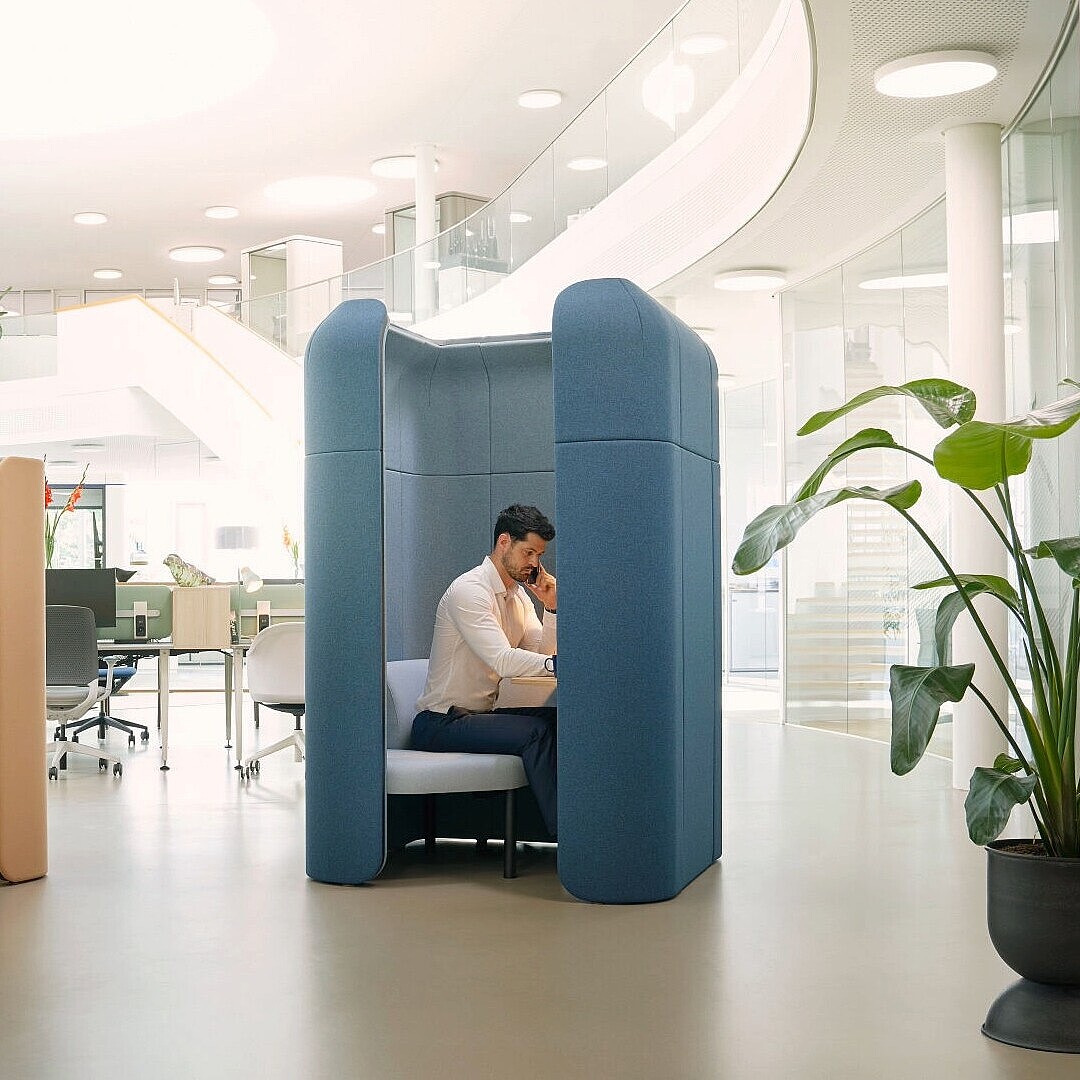 Two modular se:hive seating units in an open office space, one beige with two people seated inside and one blue with a single person seated, both designed as semi-enclosed retreats.