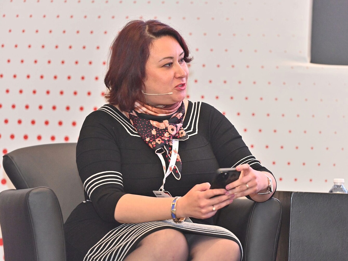 Mujer sentada con un vestido negro a rayas blancas y un pañuelo estampado, sosteniendo un smartphone en una conferencia