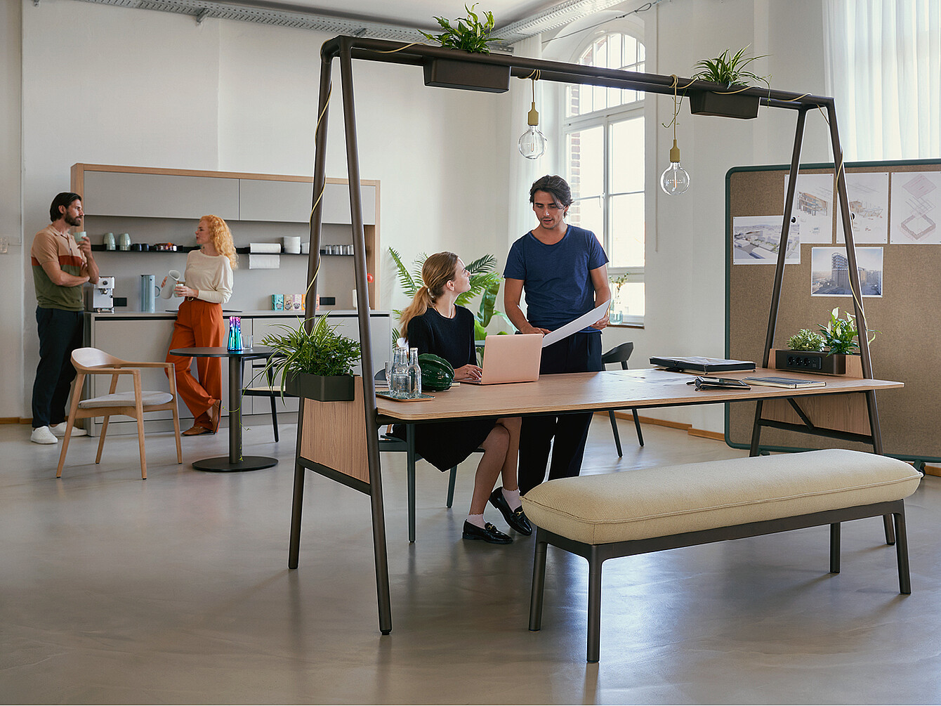Espace de travail hybride et ductile moderne où deux personnes collaborent à une table en bois avec des plantes et des suspensions lumineuses, tandis que deux autres discutent près d’une kitchenette en arrière-plan.