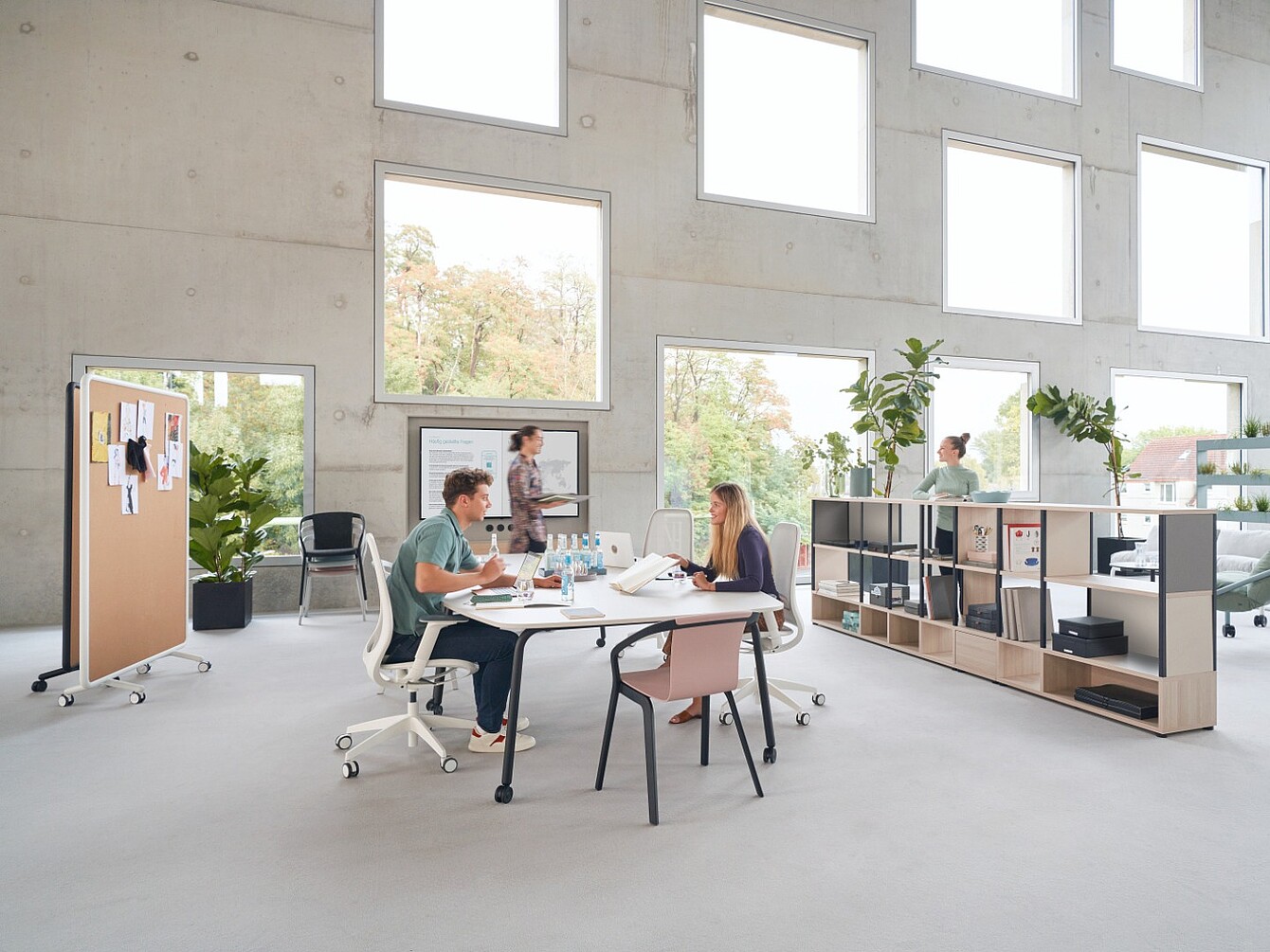 Dos personas sentadas a una mesa trabajando con papeles y ordenadores portátiles en una oficina luminosa con grandes ventanas cuadradas y una estantería