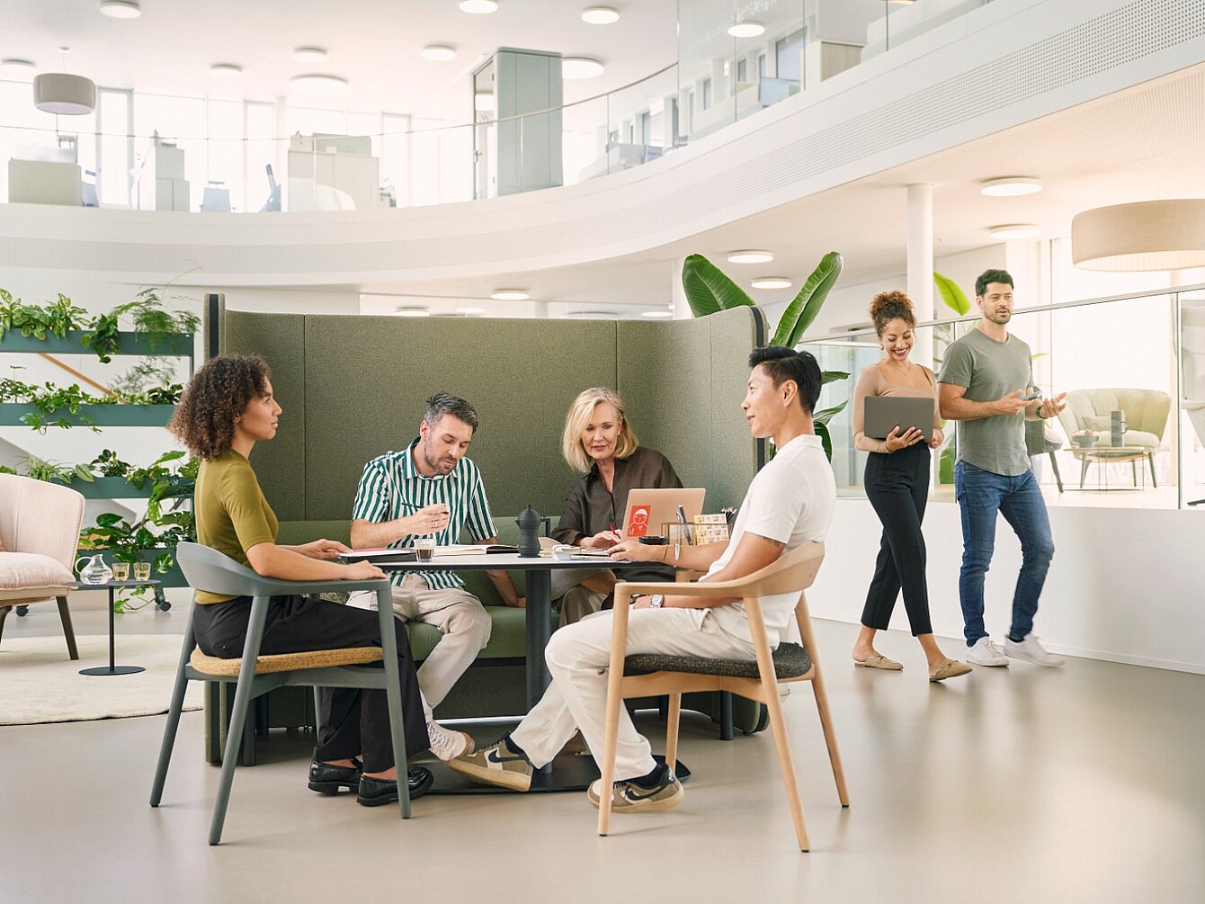 Three people sit around a round table in a modern office while two others walk past in the background.
