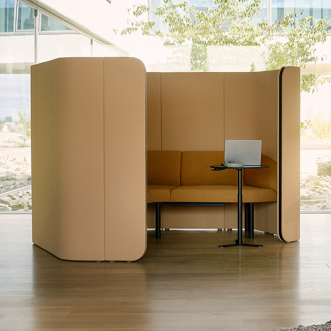 Cabina de asiento aislada con sofá, mesa pequeña y ordenador portátil en un entorno de oficina moderno y luminoso.