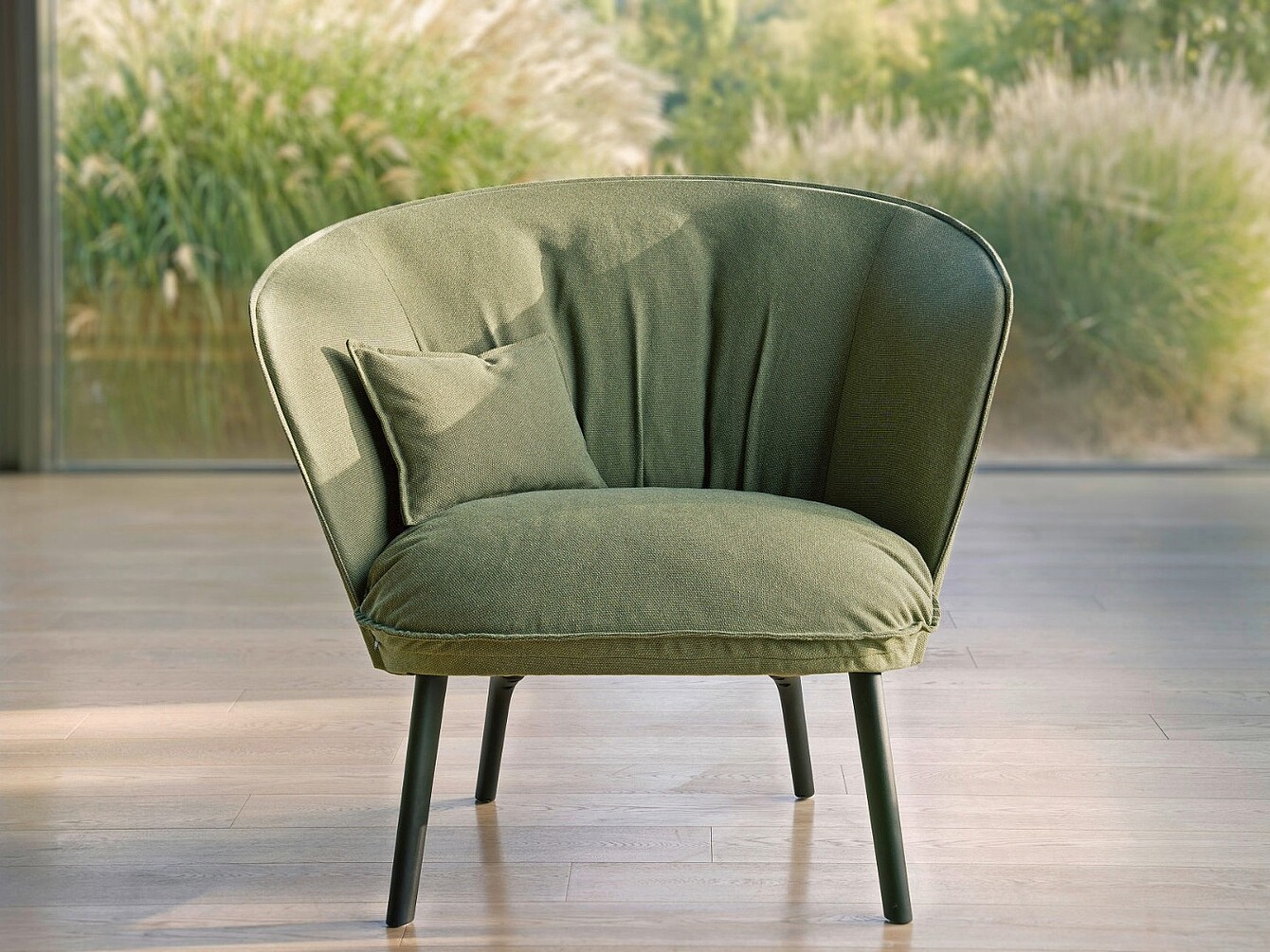 Fauteuil en tissu vert avec un coussin assorti, posé sur un sol en bois avec des plantes floues en arrière-plan