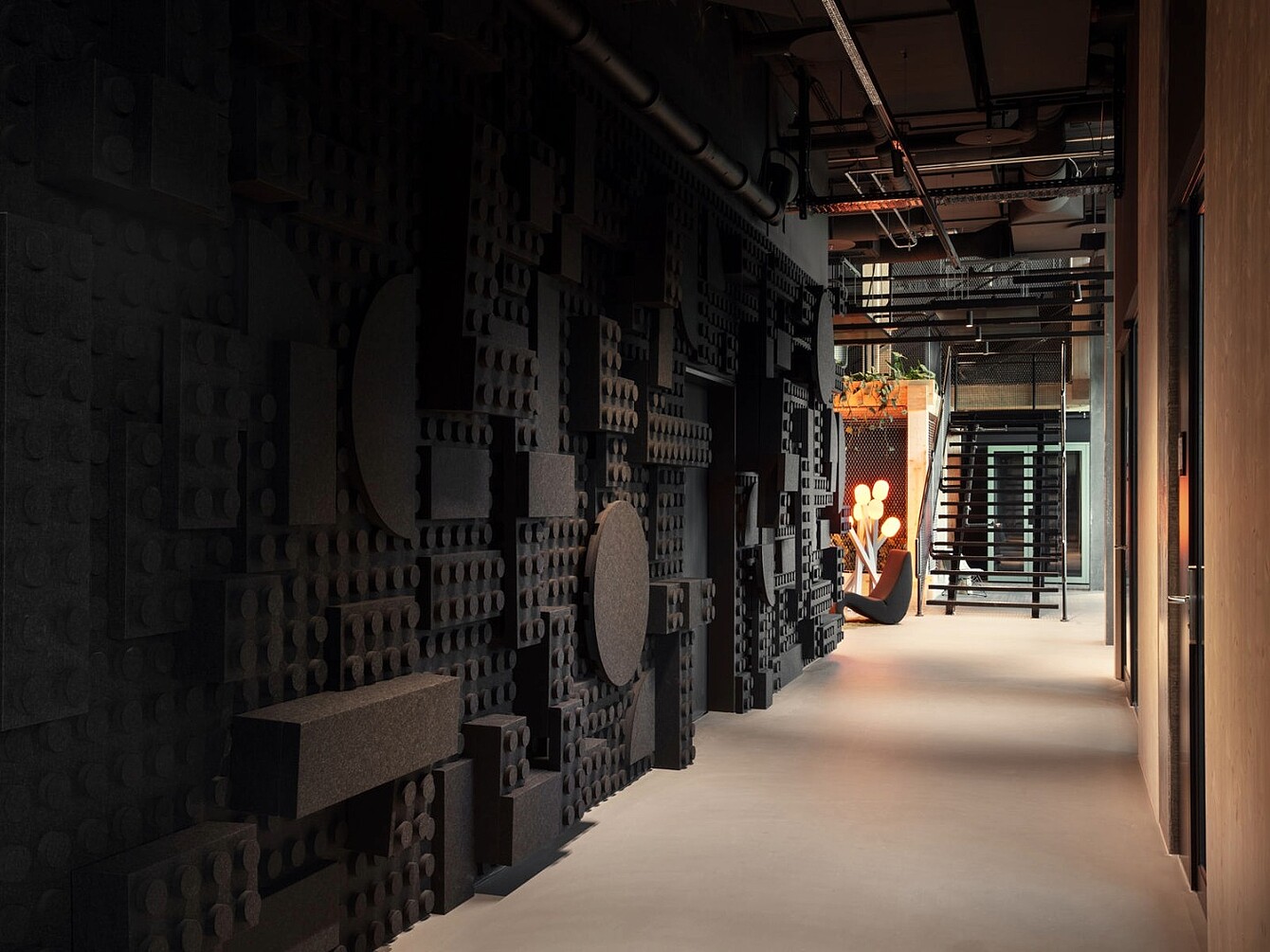 Long couloir avec un mur sombre recouvert de grands panneaux géométriques en mousse et un sol clair, menant à un coin salon avec une personne et un escalier.