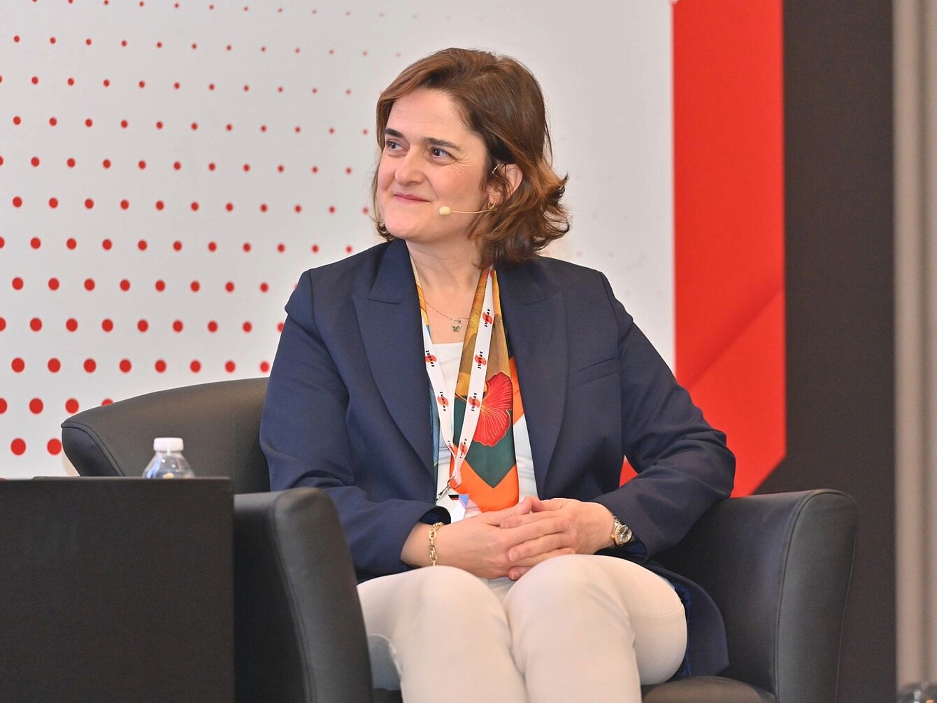 Persona sentada en un sillón negro con una chaqueta azul marino, pantalones blancos y un pañuelo de colores, con las manos entrelazadas en el regazo