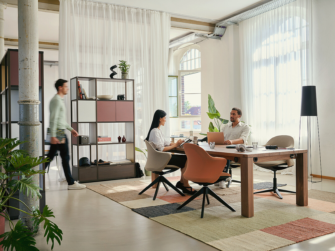 Espace de travail hybride moderne et flexible avec deux personnes assises à une table en bois utilisant un ordinateur portable et une tablette, entourées de plantes, d’étagères et de grandes fenêtres avec des rideaux transparents, soulignant la flexibilité et l’esprit communautaire.
