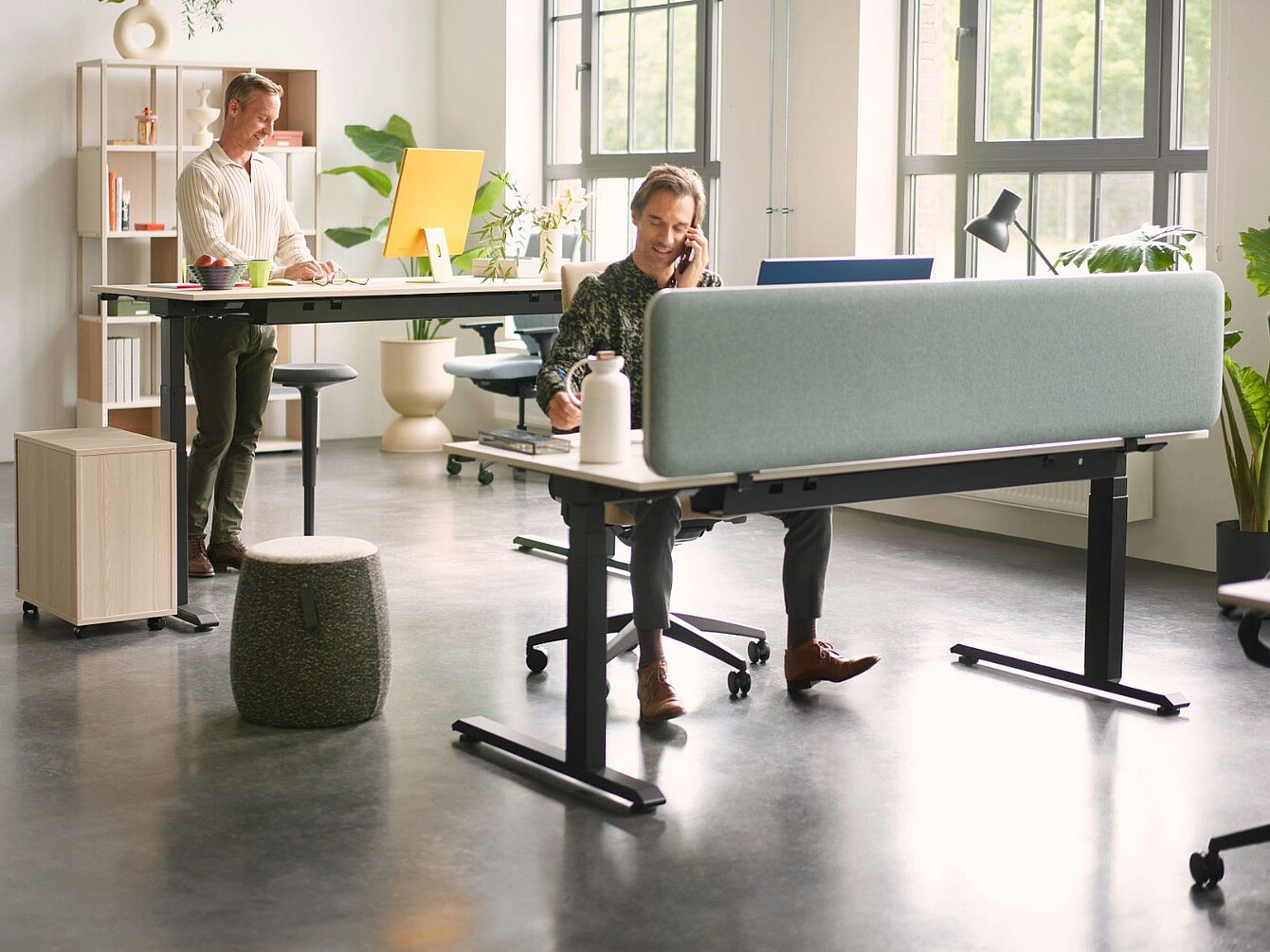Espace de bureau moderne avec deux personnes travaillant à des bureaux séparés par un écran de confidentialité bleu clair, équipé de sièges ergonomiques, d’un bureau réglable en hauteur et de lumière naturelle provenant de grandes fenêtres.