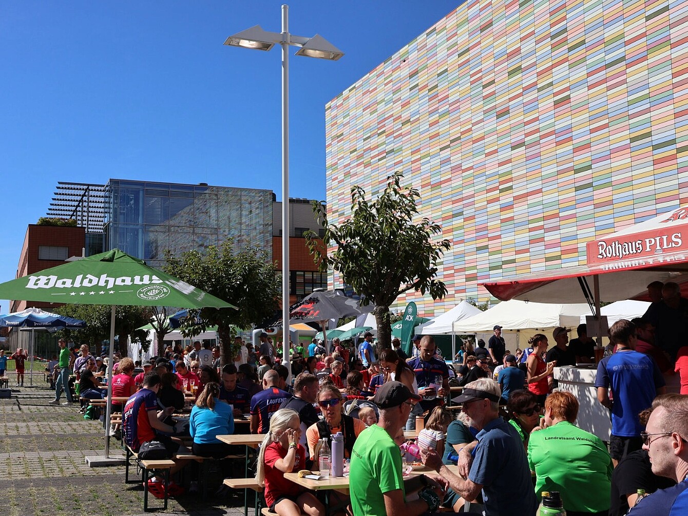 Outdoor event with groups of people sitting at picnic tables under umbrellas near a large colorful tiled building