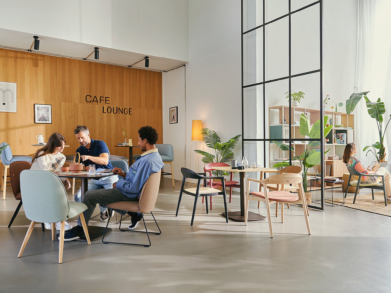 Three people sitting and talking at a table in a modern office cafe lounge with wooden walls, plants, and various seating options including armchairs and stools