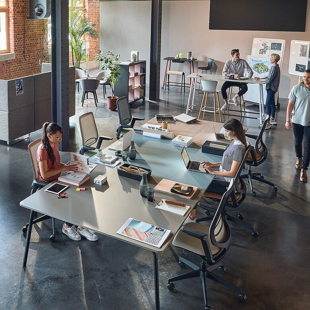 Open office space with people working at desks, exposed brick walls, large windows, and modern office furniture