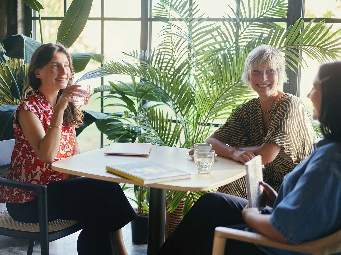 Tres personas sentadas alrededor de una mesa en un espacio de oficina luminoso con plantas grandes al fondo