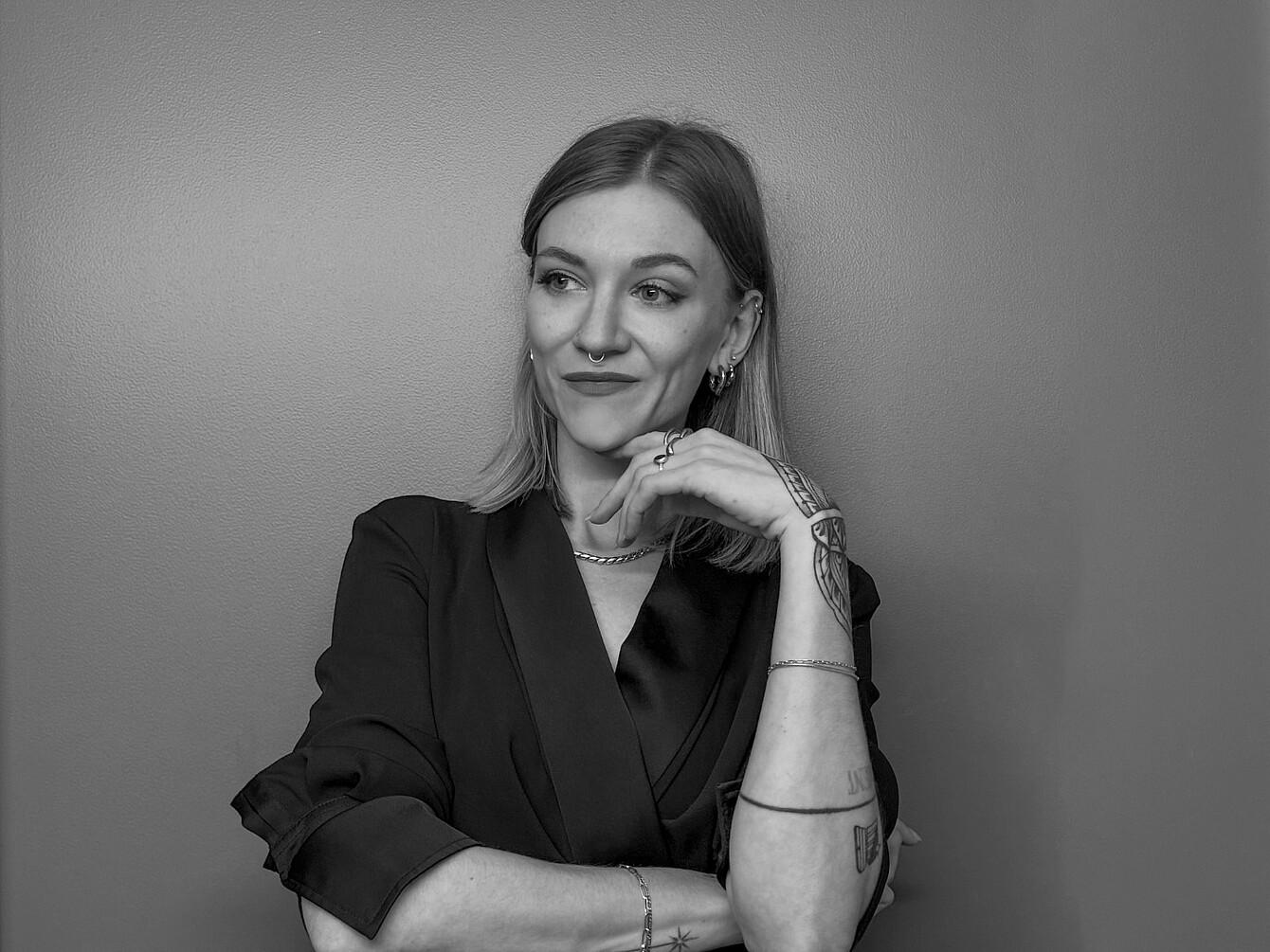 Femme aux cheveux mi-longs vêtue d’un blazer foncé, le menton posé sur la main, arborant plusieurs bracelets et tatouages sur l’avant-bras.