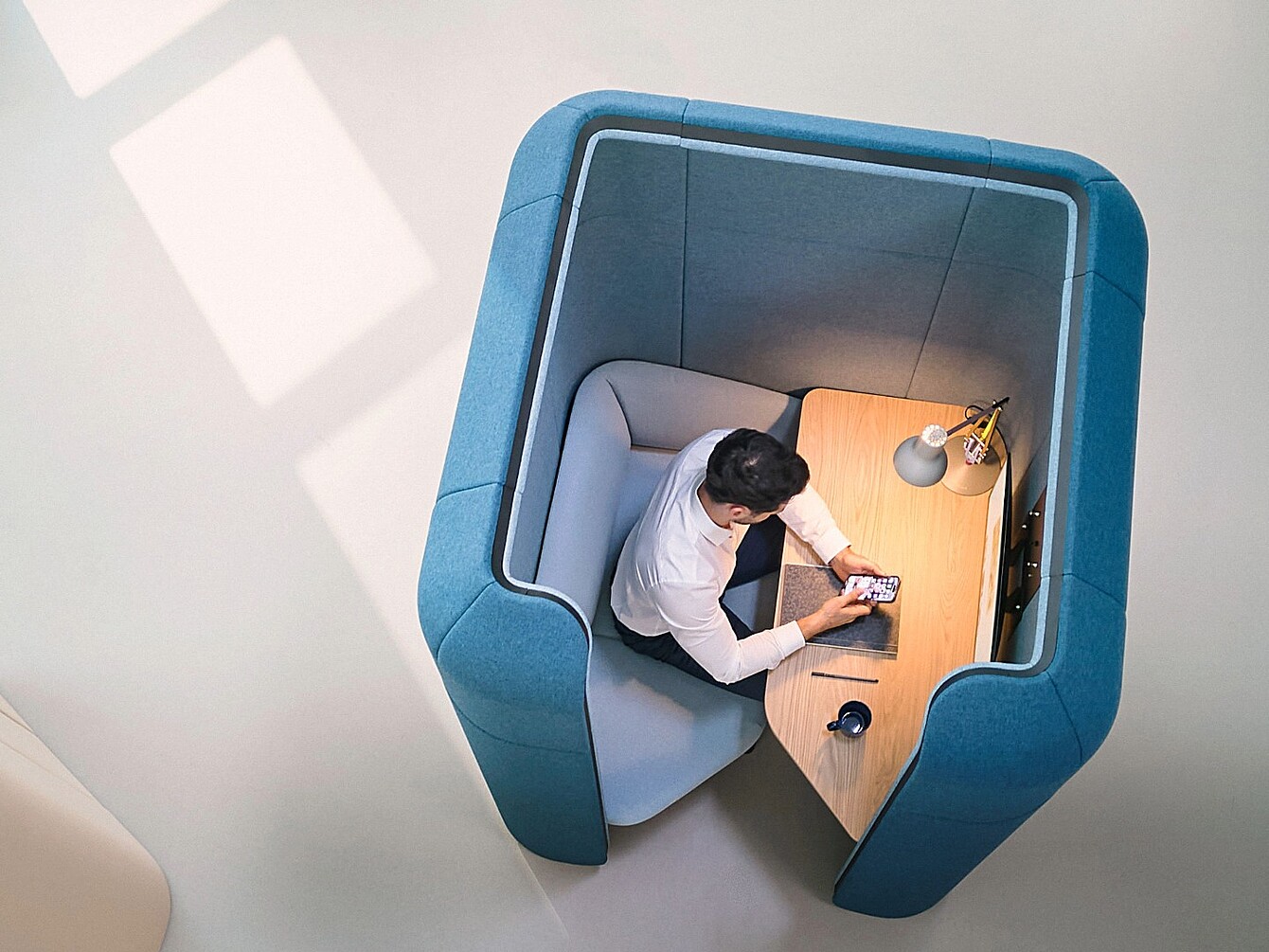 Homme assis dans un espace de travail cloisonné aux murs hauts et recouvert de tissu bleu, utilisant un smartphone sur un petit bureau en bois équipé d’une lampe et d’une tasse