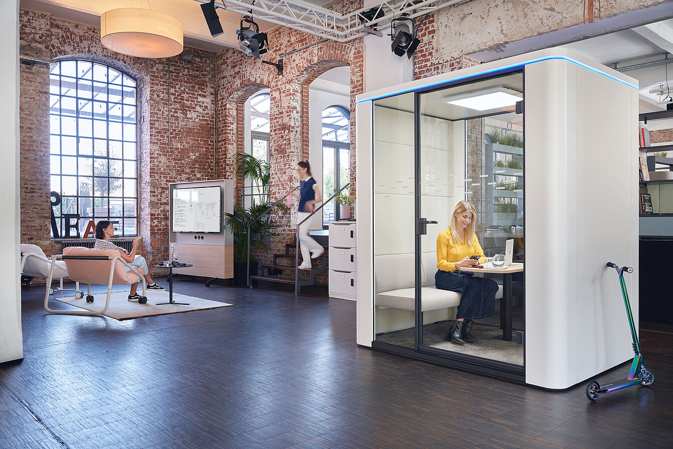 soluzione se:cube max room-in-room con una donna che utilizza il nuovo display touch all’interno, con illuminazione e ventilazione regolabili in un ufficio moderno con pareti in mattoni a vista