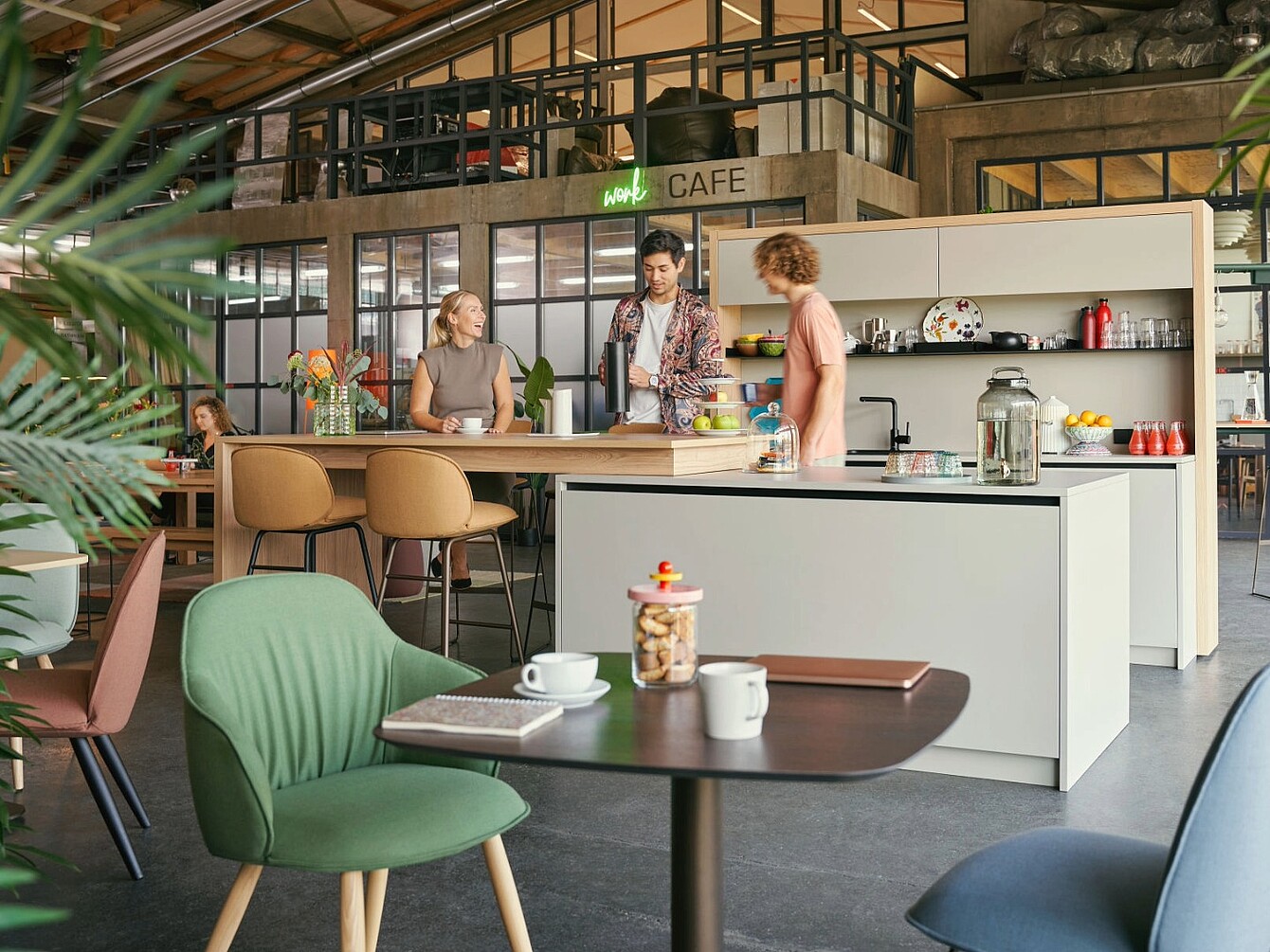 Tres personas de pie hablando alrededor de una isla de cocina en la zona de cafetería de una oficina moderna, con asientos y plantas
