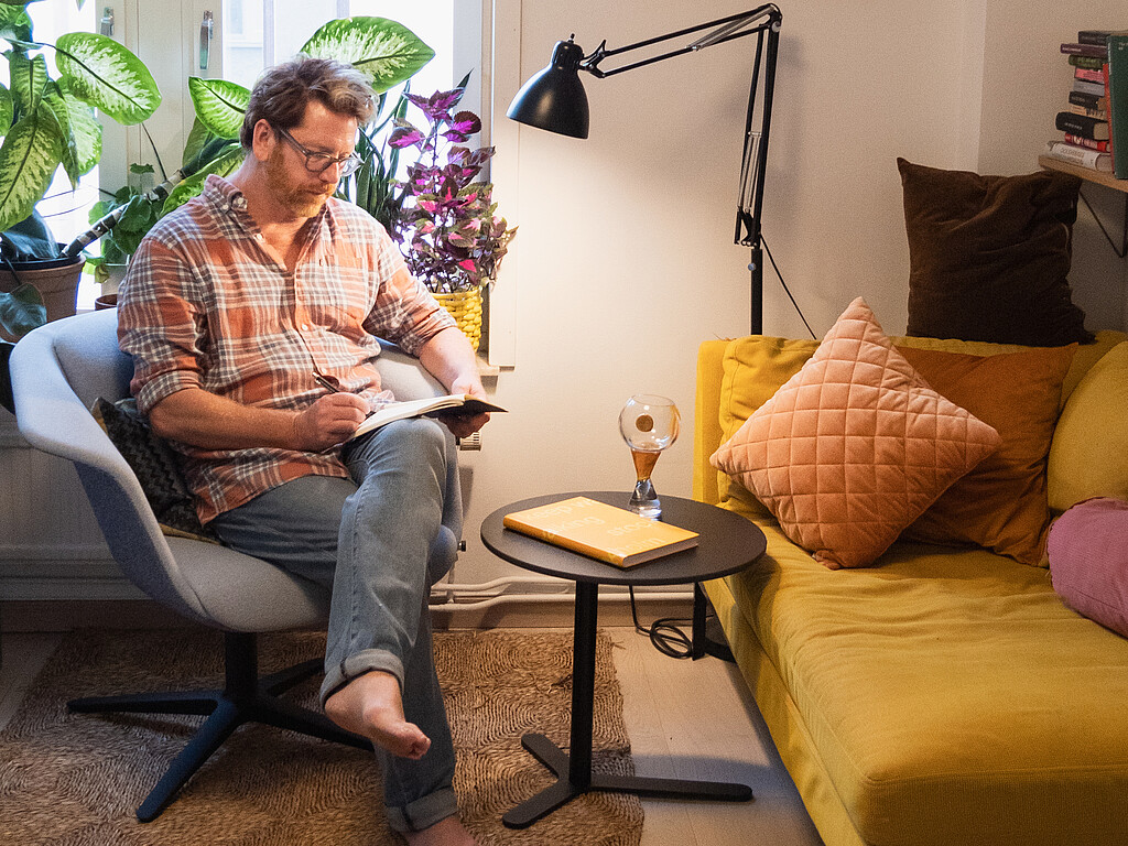 Person sitting barefoot in a grey swivel chair, writing in a notebook, surrounded by plants, next to a yellow sofa with cushions and a black floor lamp.