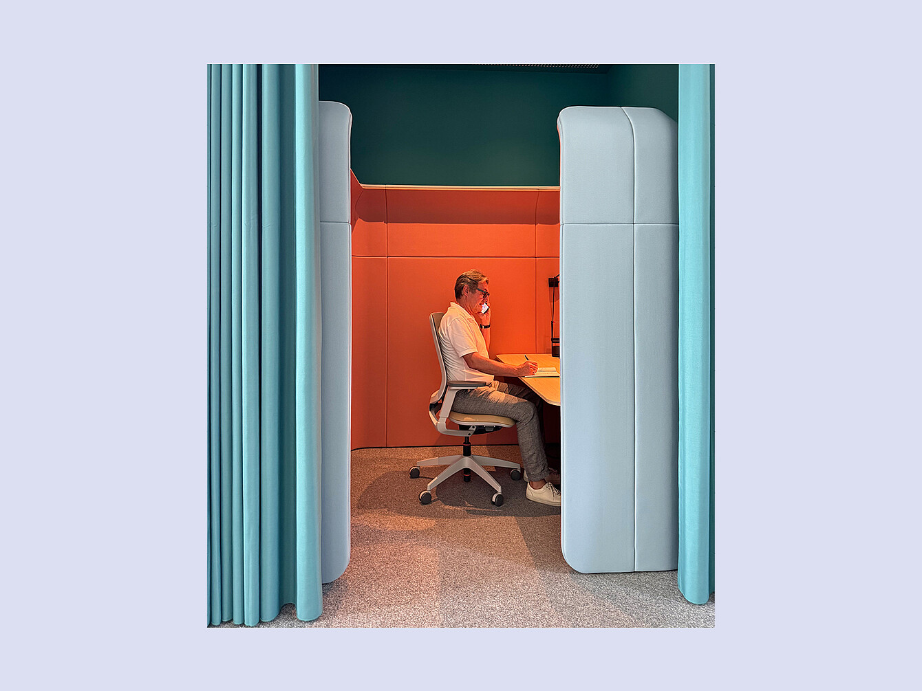 Personne assise sur une chaise de bureau, travaillant à un bureau dans un petit espace de travail fermé, avec des murs capitonnés et un rideau sur un côté