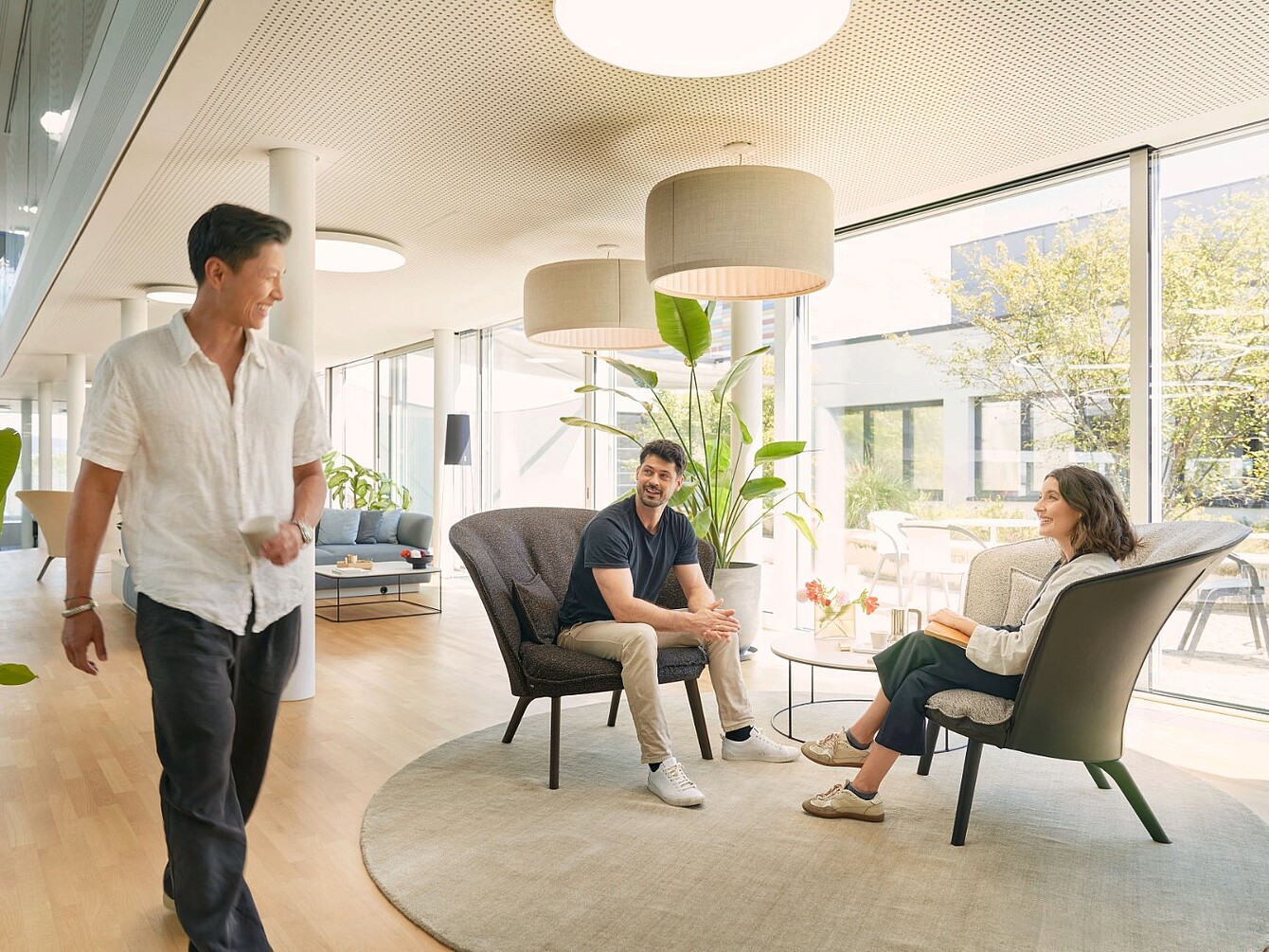 Trois personnes dans un salon de bureau moderne doté de grandes fenêtres, assises et debout autour d’un tapis rond et de fauteuils