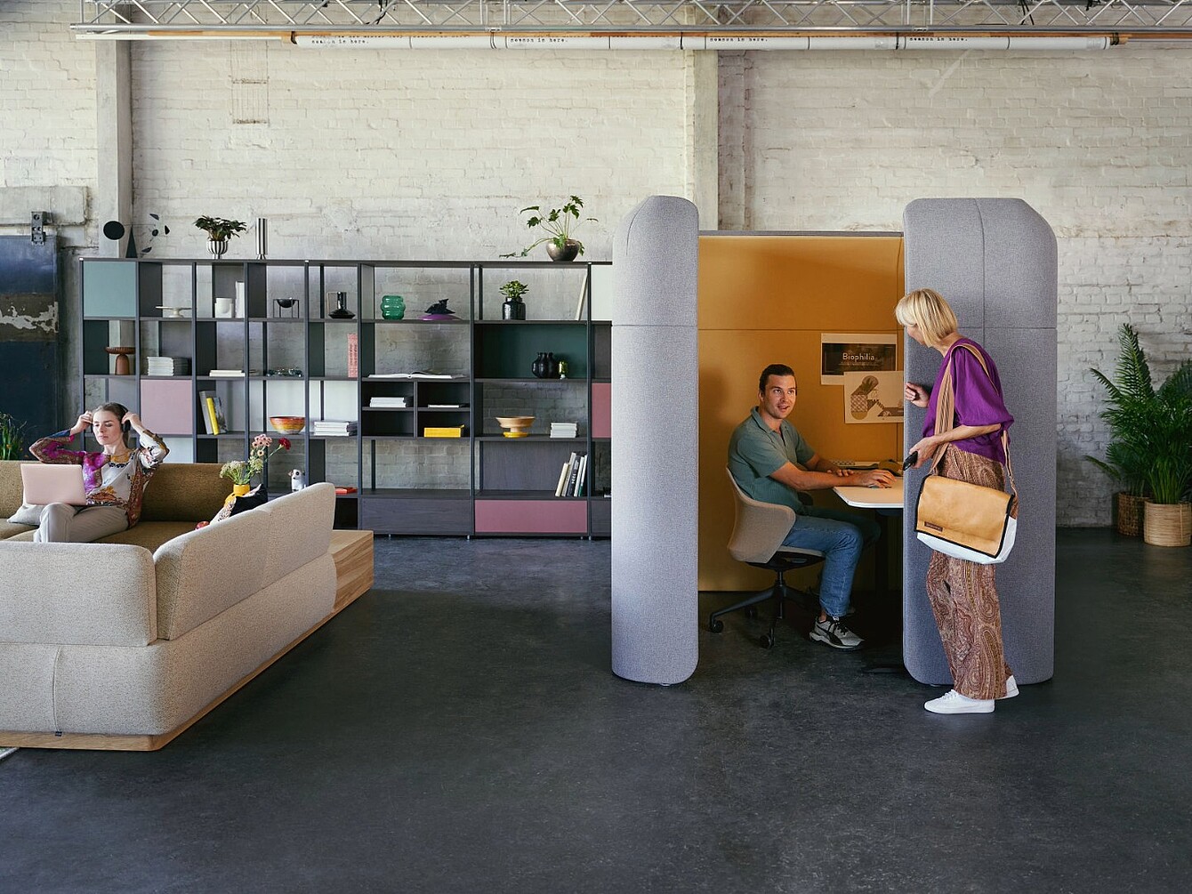 Un homme assis à l’intérieur d’un petit espace de travail clos, avec de hauts murs gris et un intérieur orange, utilisant un ordinateur portable, tandis qu’une femme se tient à l’extérieur de l’espace et lui parle, dans un bureau moderne avec un canapé et une étagère en arrière-plan.