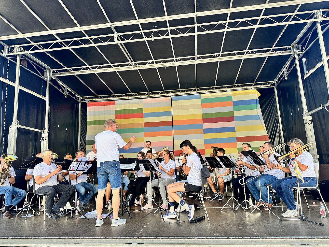 Conductor of the Sedus Werksmusik in white shirt and shorts leads seated musicians playing brass and woodwind instruments on stage with colorful striped backdrop.