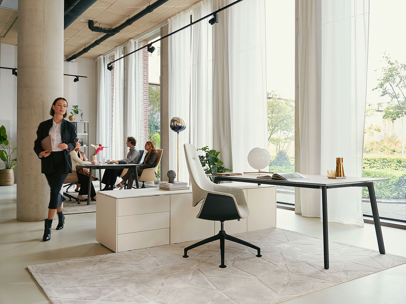 Espace de bureau moderne avec de grandes fenêtres, des rideaux blancs, un bureau avec une chaise pivotante Sedus se:mission blanche et des personnes assises et marchant