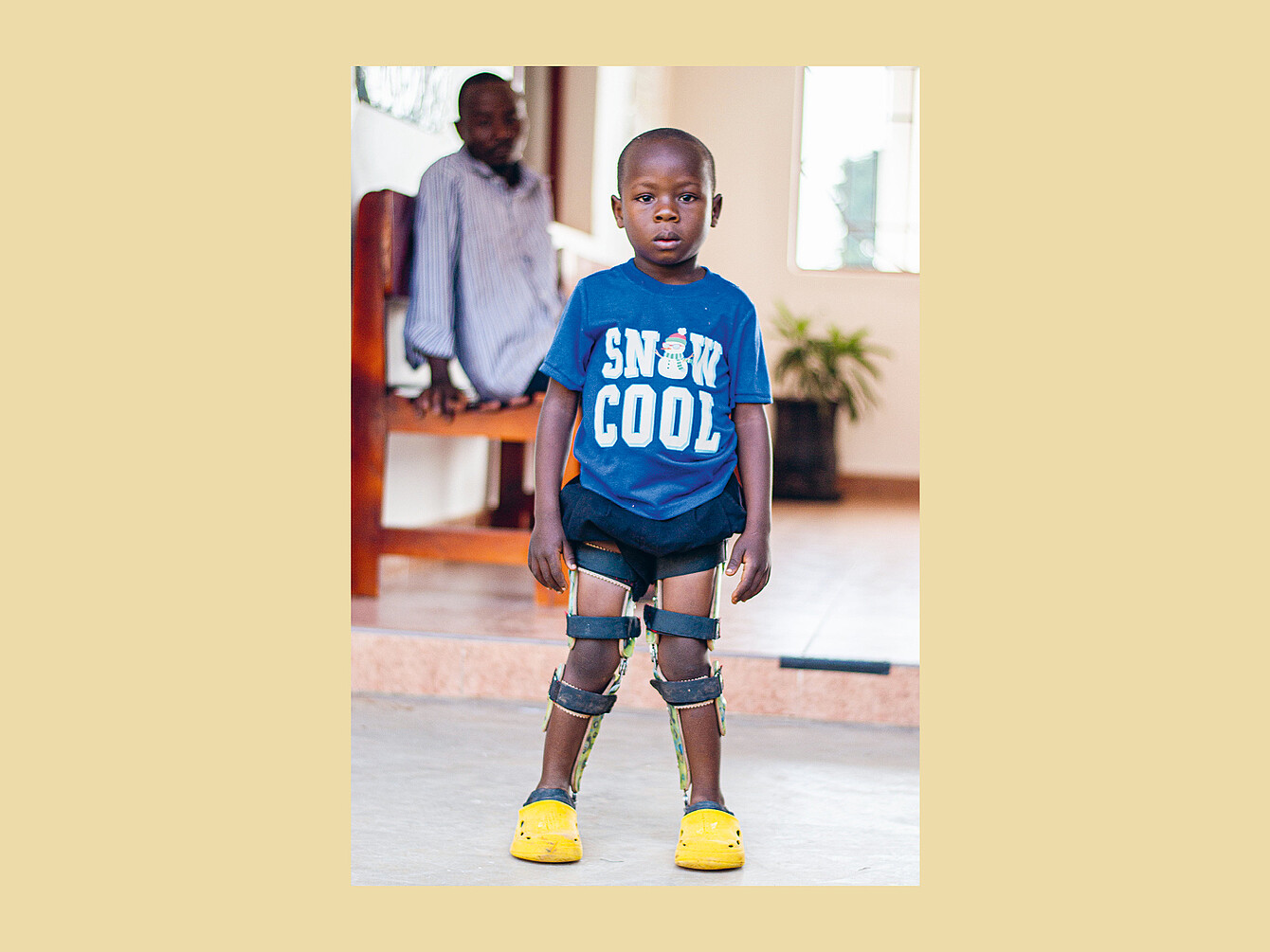 Child standing indoors wearing a blue T-shirt with 'SNOW COOL' text and snowman graphic, black shorts, leg braces, and yellow clogs.
