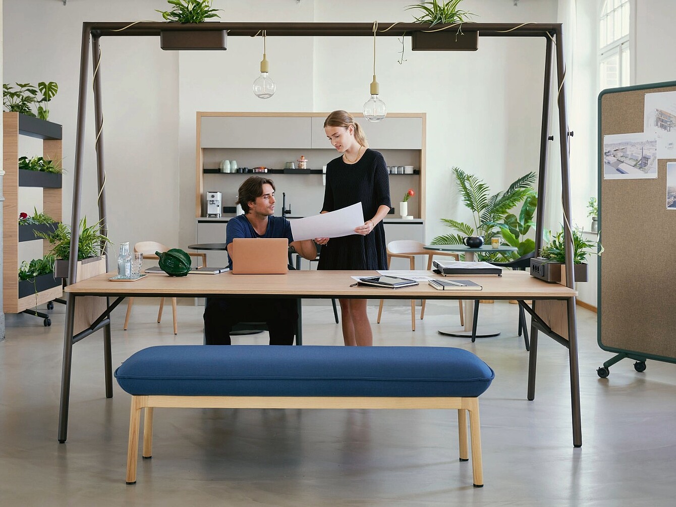 The picture shows a modern workspace with a large desk where two people are looking at documents together. Above the desk is an open-frame structure with in-tegrated plants and lighting, which visually defines the workspace. The light-coloured