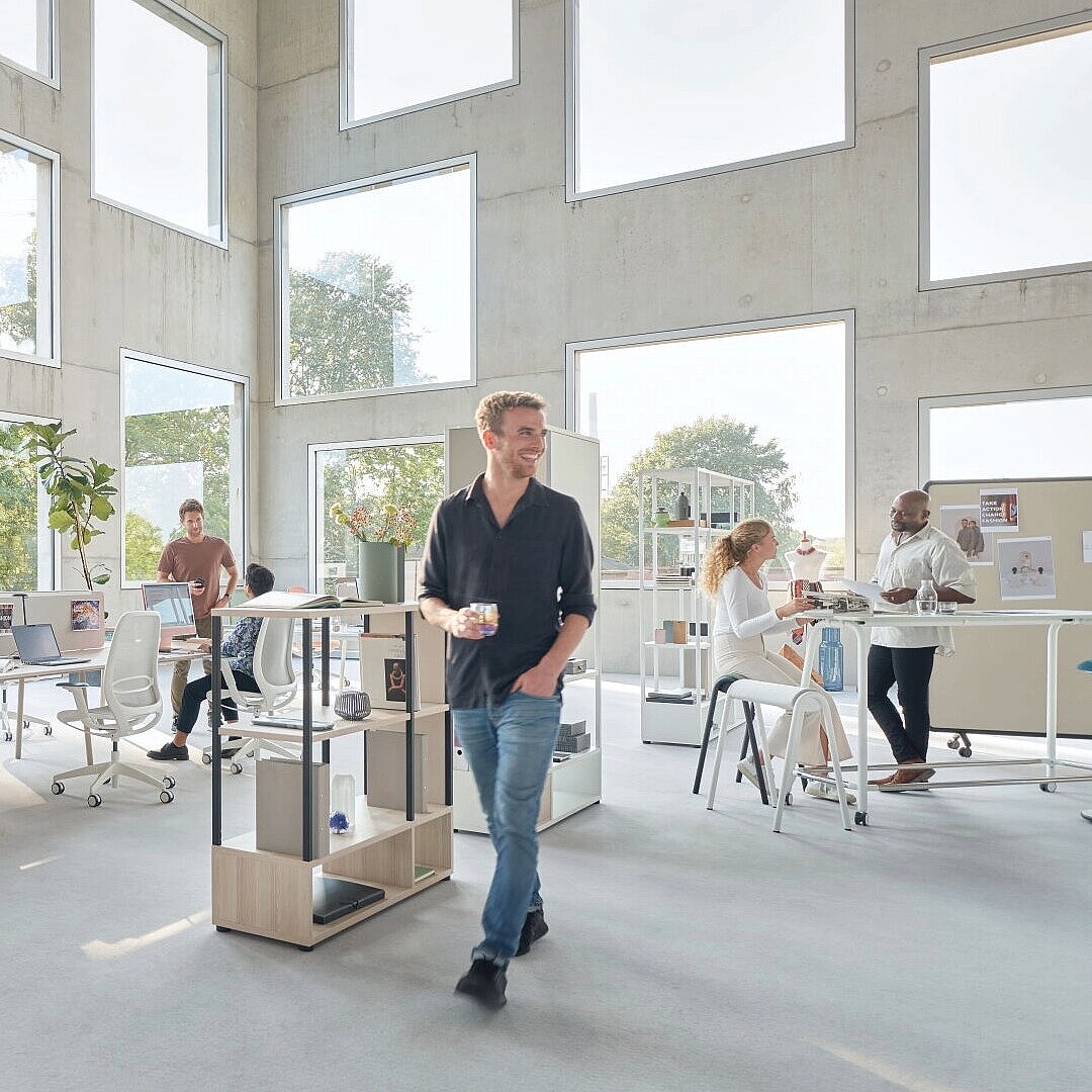 Open-plan office with multiple people working and interacting at desks and standing in a bright space with large windows and plants
