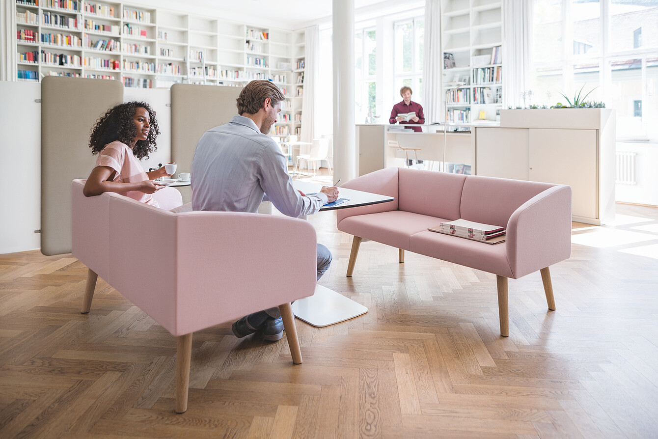 Bright library-inspired workspace with pink sofas, acoustic screens, bookshelves and people working quietly.