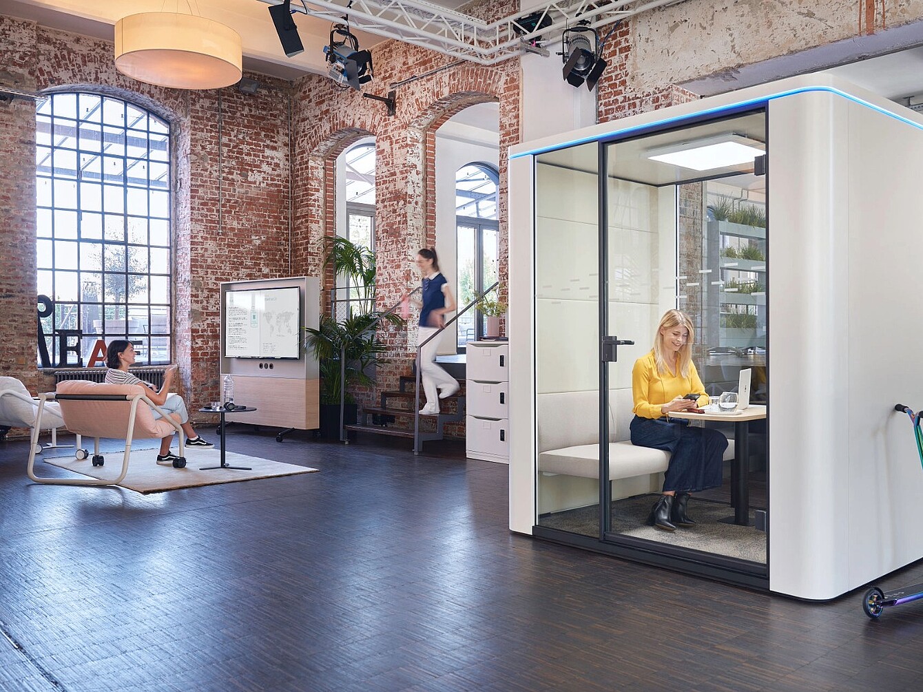 Espace de bureau moderne avec des murs en briques apparentes et de grandes fenêtres, représentant une femme assise à l’intérieur d’une cabine acoustique vitrée travaillant sur un ordinateur portable et deux autres femmes dans l’espace ouvert, l’une assise sur une chaise et l’autre montant les escaliers