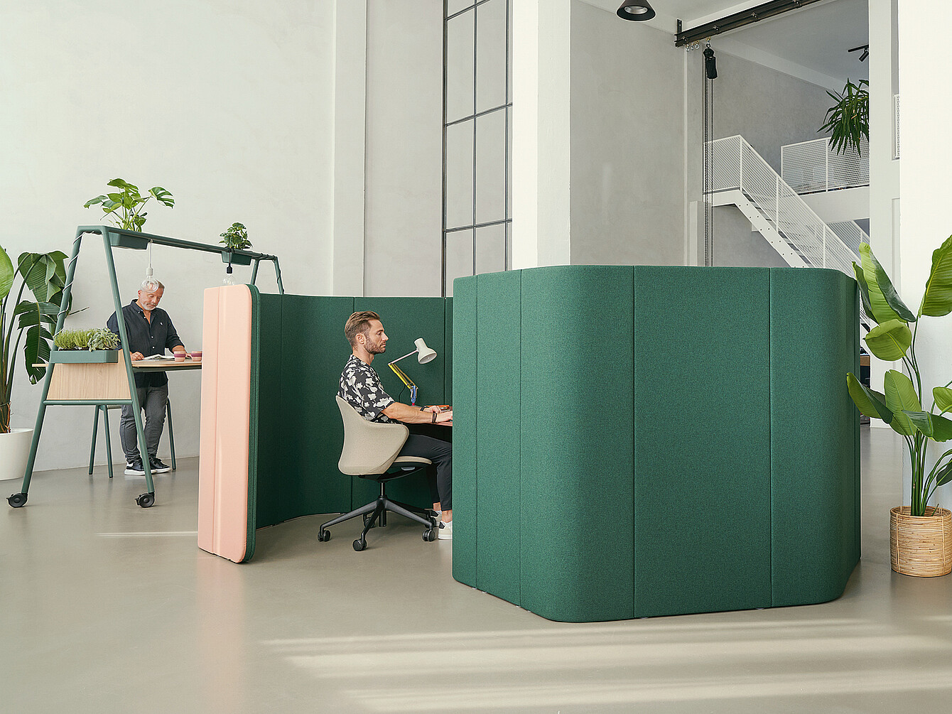 Dos puestos de trabajo en una oficina moderna con mamparas acústicas verdes. Una persona está sentada en un escritorio con una lámpara, mientras que otra está de pie junto a una mesa más alta con plantas.