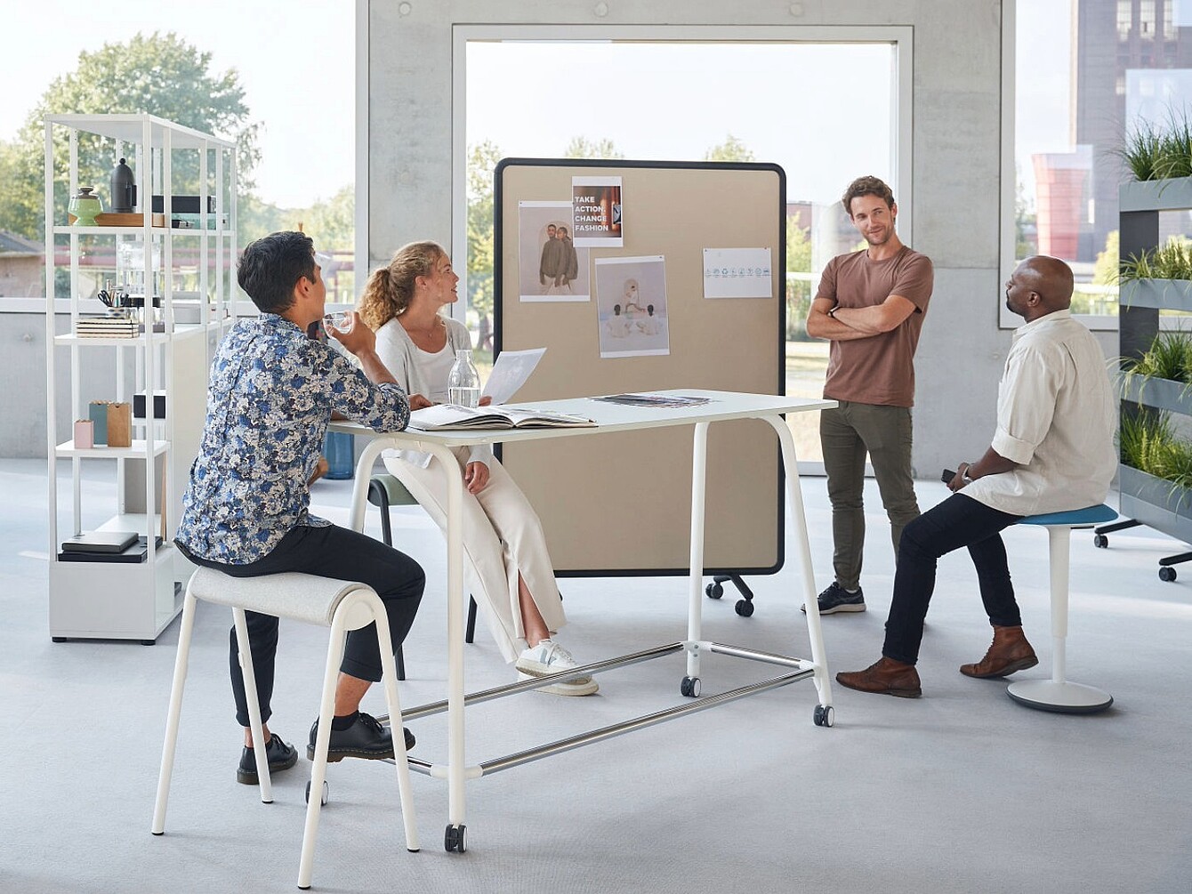 The picture shows a modern, open-plan office area where several people are work-ing together at a high table and discussing documents. Movable partitions, shelving and plants help organise the space and create flexible areas for teamwork and col-laboration. Large windows let in plenty of natural light, creating a bright and wel-coming working environment.