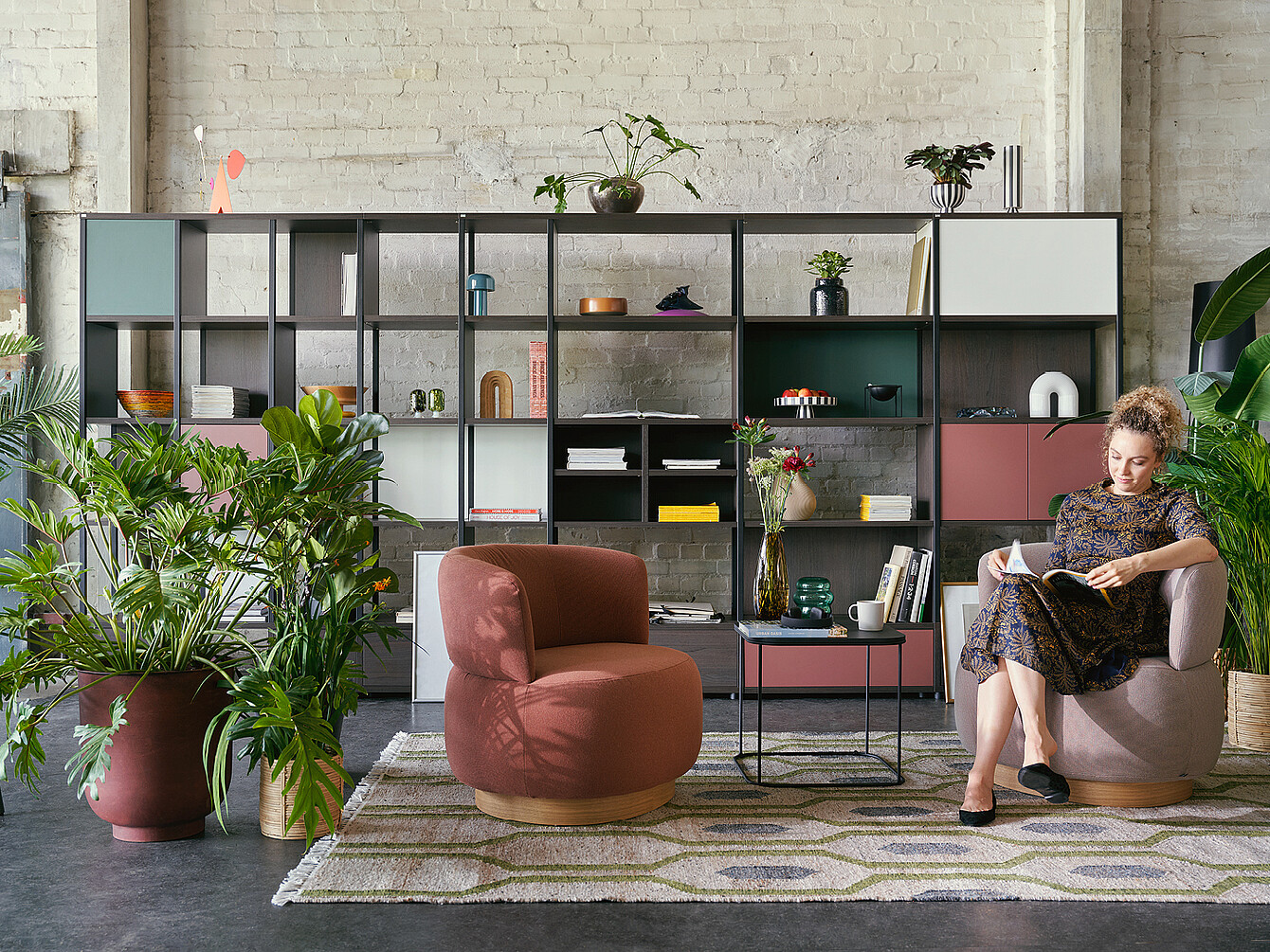 Personne lisant dans un cadre élégant du Work Café Club avec des fauteuils modernes, un tapis géométrique, des étagères modulaires avec des plantes et des éléments décoratifs, et un mur en briques apparentes