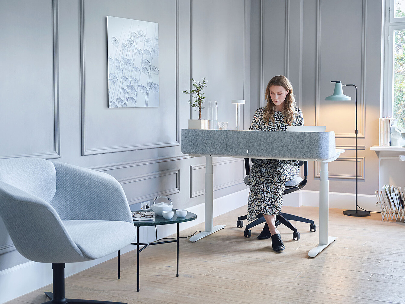 Femme en robe à fleurs assise à un bureau Sedus blanc moderne dans une pièce gris clair avec un fauteuil bleu et un lampadaire