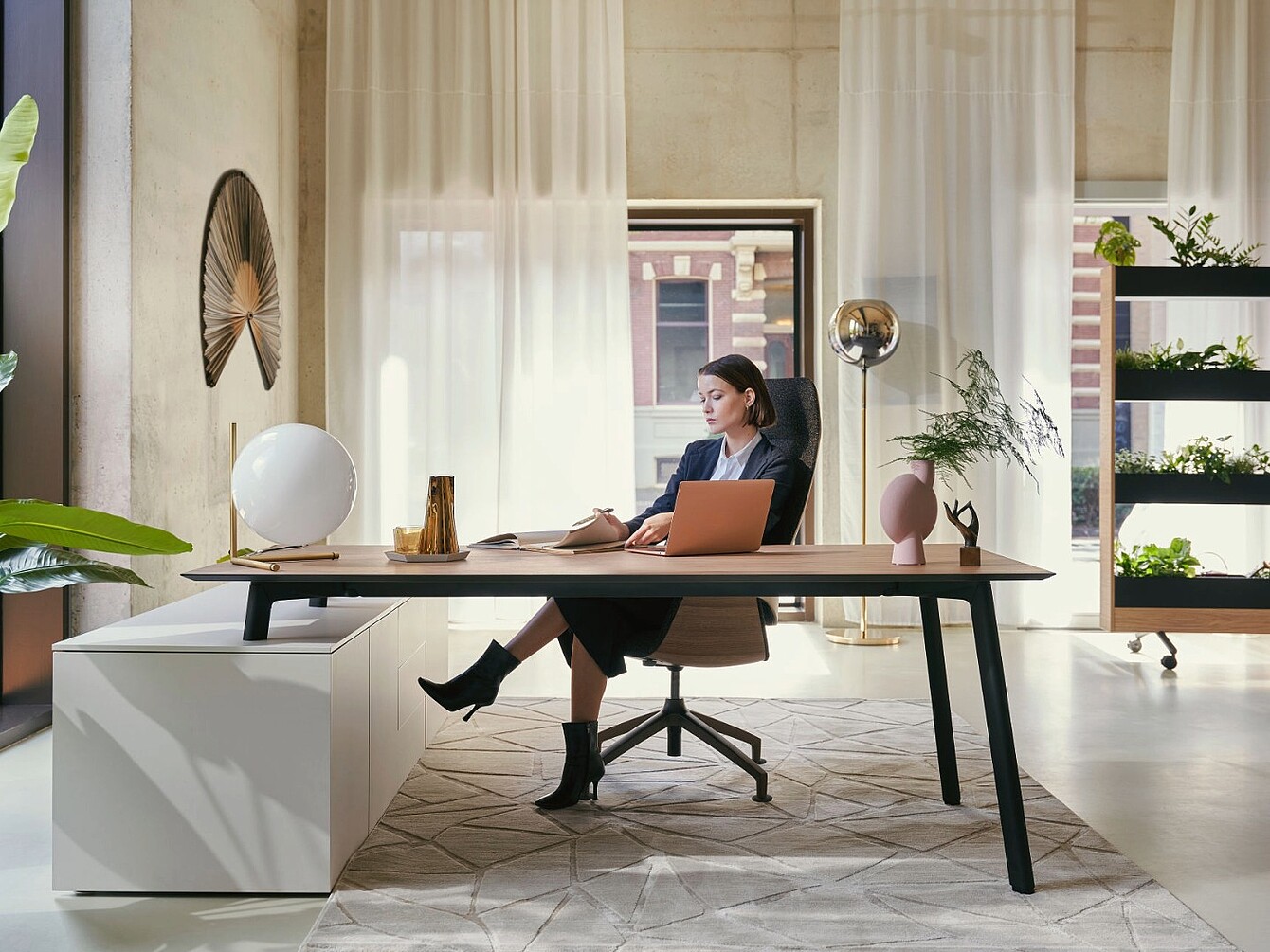 Mujer sentada en un moderno escritorio de oficina con un ordenador portátil y un cuaderno, en una habitación luminosa con cortinas transparentes, plantas y objetos decorativos