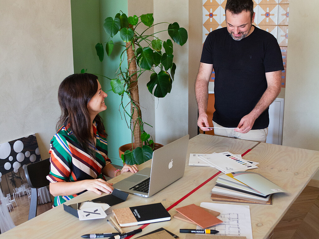 Two people working together at a wooden table with a laptop, documents, and notebooks in a bright room with a large plant.