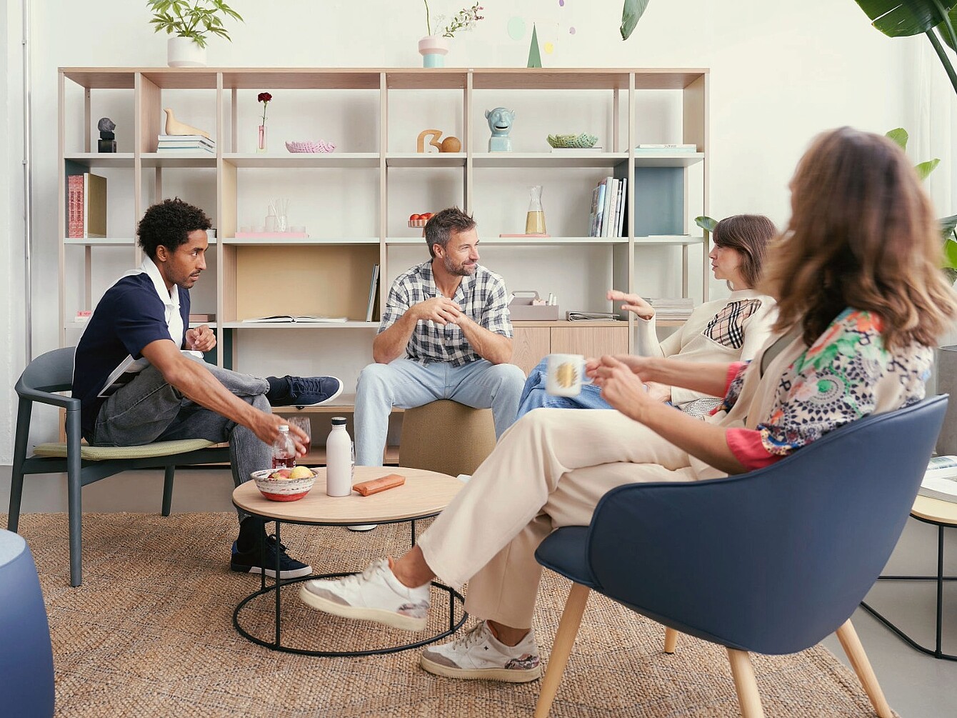 Four people sitting in a lounge area with modern chairs and a low round table in front of a large shelving unit with decorative objects and books
