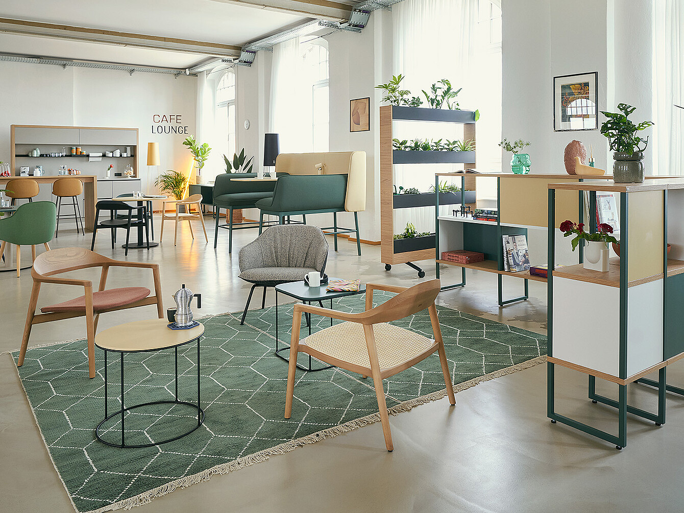 Smart Café moderno in un ufficio con sedute lounge, tappeto verde, tavoli in legno, piante e un’area lounge che unisce spazi di lavoro e di relax.
