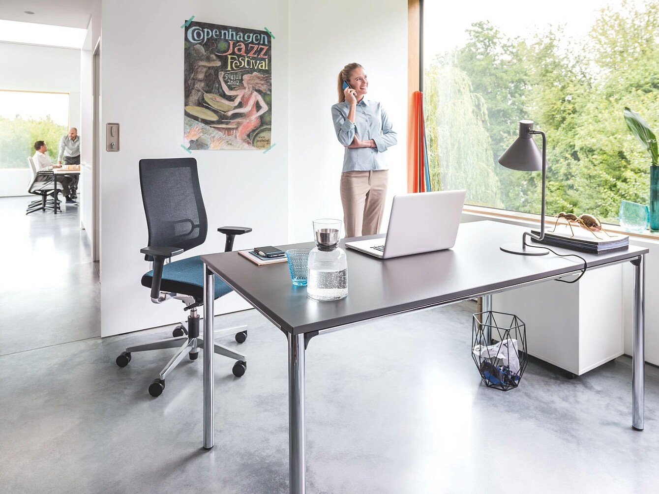Modern kantoor met een zwarte ergonomische stoel van gaas, een bureau met een laptop, een glazen waterfles, een lamp en een vrouw die bij een groot raam staat te telefoneren