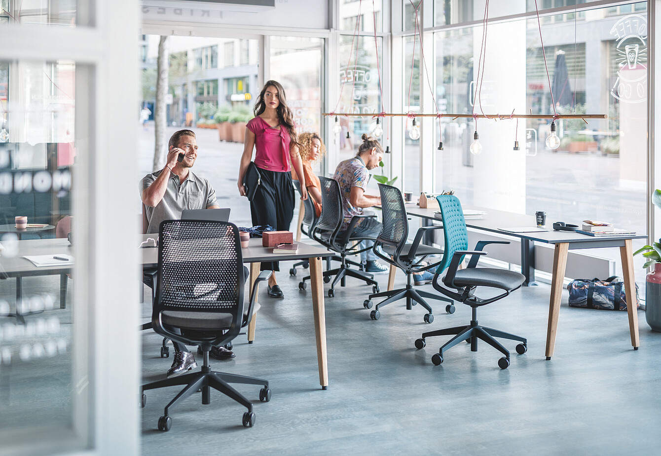 Trois personnes dans un bureau moderne doté de grandes baies vitrées et de sièges de travail en résille, autour d’une table en bois