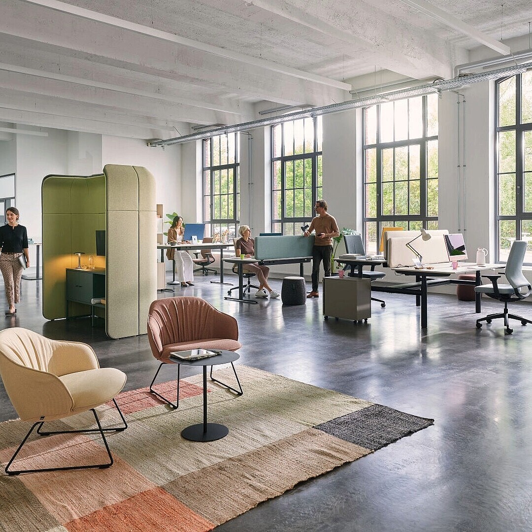 Open-plan office with large windows, multiple desks, and sound-absorbing booths where people are working or sitting